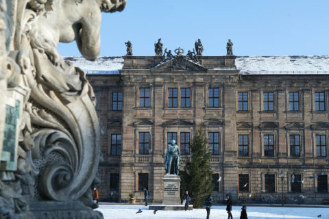 Das Bild ist ein Ausschnitt des Erlanger Schlossplatzes. Es ist eine Winteraufnahme; der Himmel ist strahlend blau und es liegt ein wenig Schnee. Auf dem Foto ist das Erlanger Schloss zu sehen, heutiger Sitz der Universität. Vor dem Schloss steht das Markgrafendenkmal, es zeigt den Universitätsgründer, Markgraf Friedrich von Brandenburg-Bayreuth. Zwischen Denkmal und Schloss steht eine sehr große dunkelgrüne Tanne.