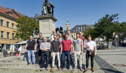 Auf dem Bild sind 11 Erwachsene zu sehen, die auf dem zentralen Platz Schweinfurts stehen. Die Gruppe steht direkt vor einer Bronzestatue, im Hintergrund ist der Glockenturm einer Kirche zu sehen. Alle tragen legere Kleidung und lächeln in die Kamera.